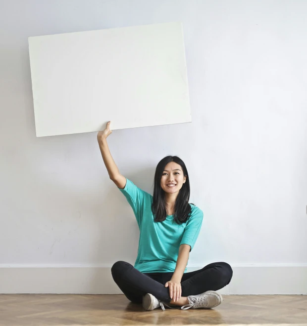 a woman sitting on the floor holding up a sign, a minimalist painting, pexels contest winner, whiteboard, half asian, waving, square