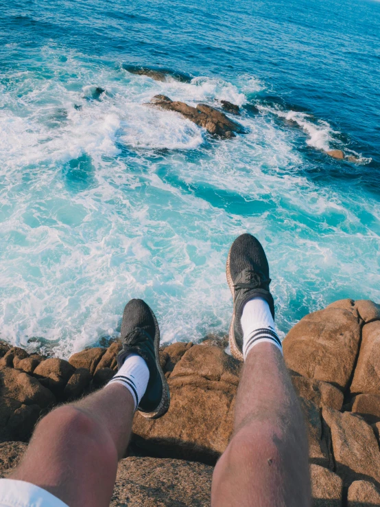 a man sitting on top of a rocky cliff next to the ocean, legs taking your pov, teal aesthetic, high soles, 2019 trending photo