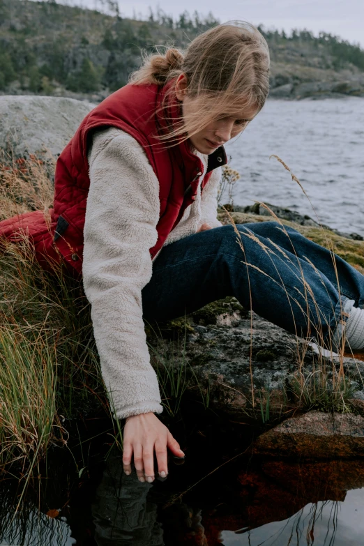 a woman sitting on a rock next to a body of water, by Christen Dalsgaard, wearing a red gilet, touching her clothes, subsurface scandering, crawling on the ground