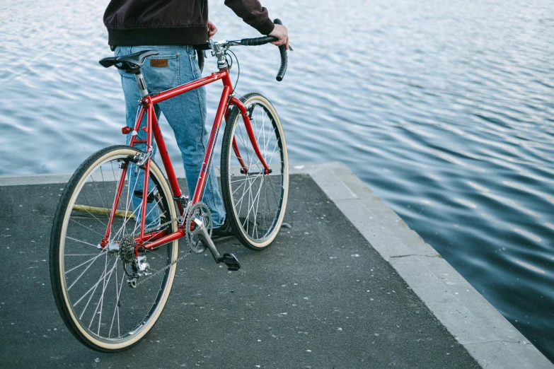 a man standing next to a red bike next to a body of water, pexels contest winner, legs visible, paved, thumbnail, rectangle