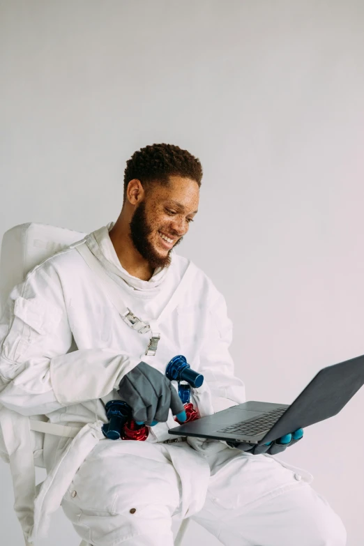 a man sitting in a chair with a laptop, inspired by John Gibson, pexels contest winner, afrofuturism, with a lab coat, nasa eva suit, smiling male, dirty linen robes