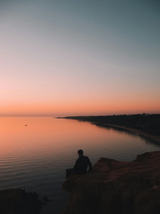 a person sitting on a rock next to a body of water, a picture, unsplash contest winner, pastel orange sunset, looking onto the horizon, slightly pixelated, wide high angle view