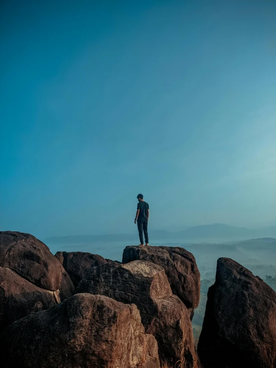 a man standing on top of a large rock, pexels contest winner, gazing off into the horizon, blue, boulders, slightly pixelated