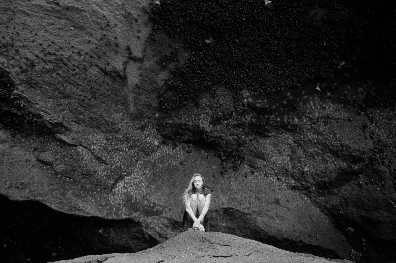 a black and white photo of a woman sitting on a rock, unsplash, minimalism, black sand, lost in a cave, ryan mcginley, wideangle portrait