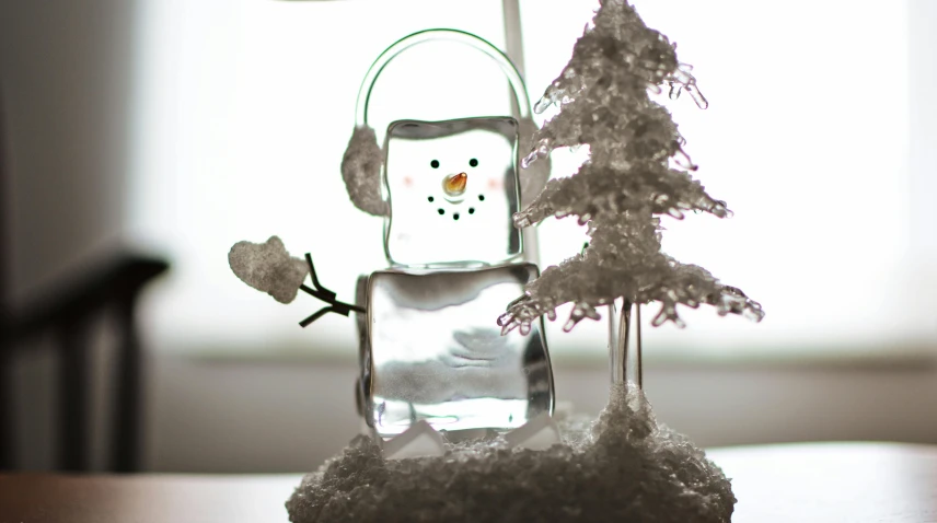 a snowman sitting on top of a table next to a christmas tree, by Sam Havadtoy, pexels, process art, person made out of glass, square, fan favorite, closeup of an adorable