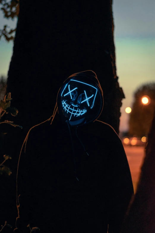 a person wearing a neon mask standing next to a tree, pexels contest winner, wearing a dark hood, crooked smile, hacker, dark hat