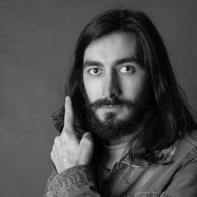 a black and white photo of a man with long hair, an album cover, by Clifford Ross, pixabay, photorealism, with a full black beard, renzo piano, color studio portrait, hand on his cheek