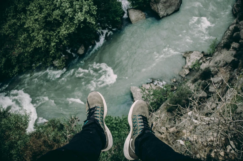 a person standing on top of a cliff next to a river, pexels contest winner, sneakers, looking up at camera, lush vista, sitting down casually