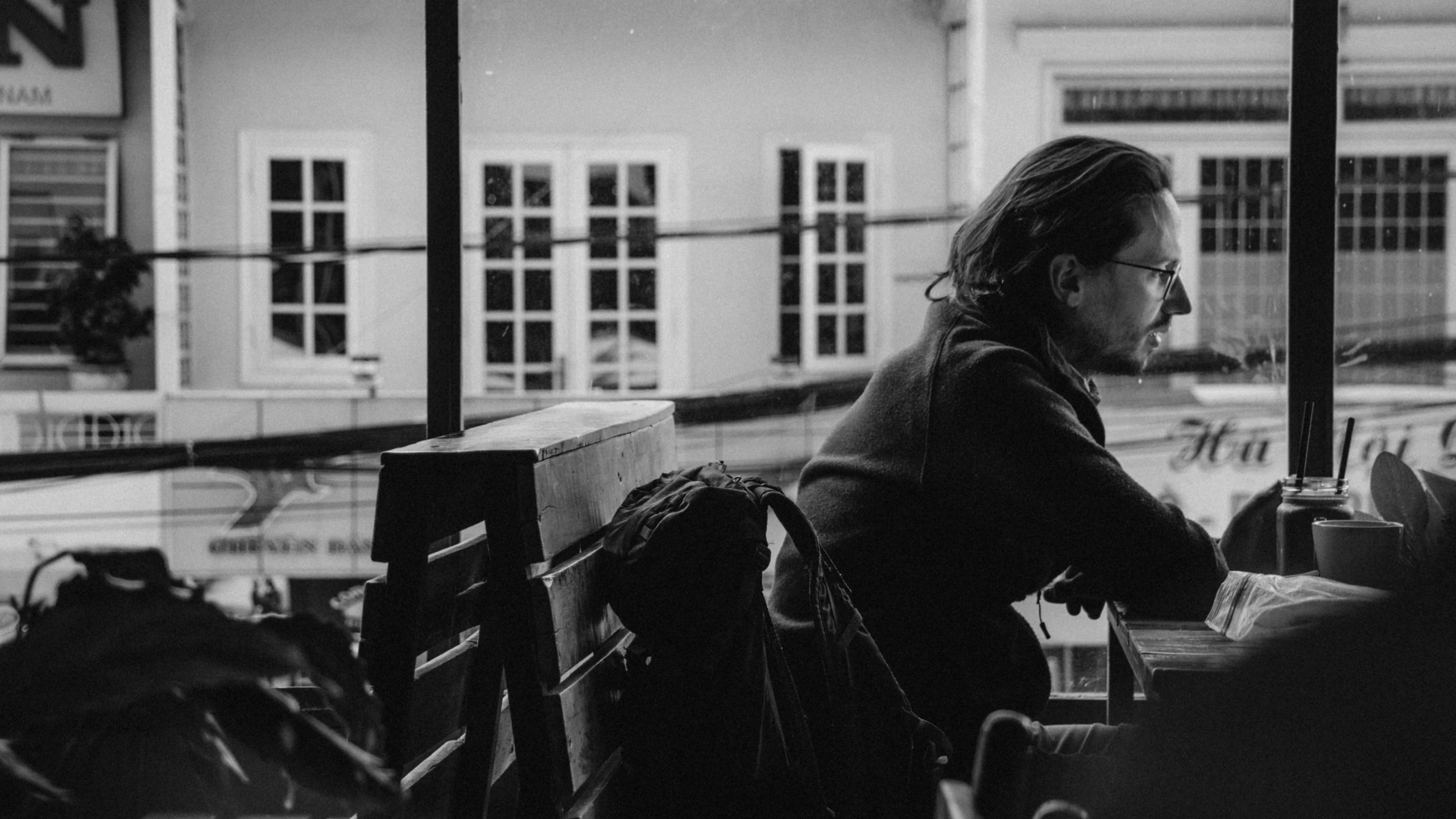 a man sitting at a table in front of a window, a black and white photo, by Emma Andijewska, hozier, raining outside the cafe, student, jovana rikalo