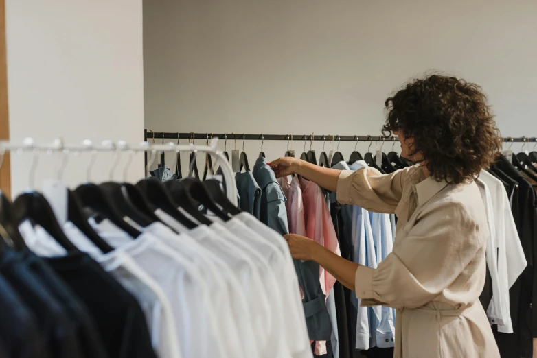 a woman standing in front of a rack of clothes, by Nicolette Macnamara, trending on pexels, inspect in inventory image, profile image, thumbnail, looking across the shoulder