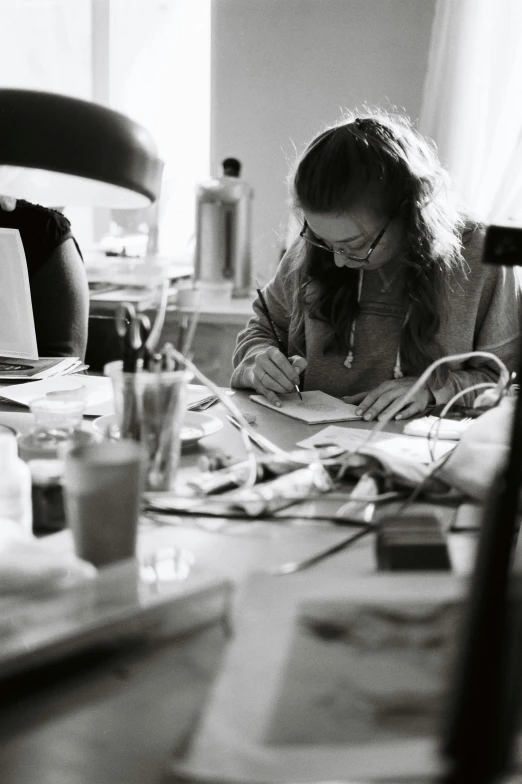 a woman sitting at a table writing on a piece of paper, process art, taken in the 2000s, studious, promo image, abigail larson