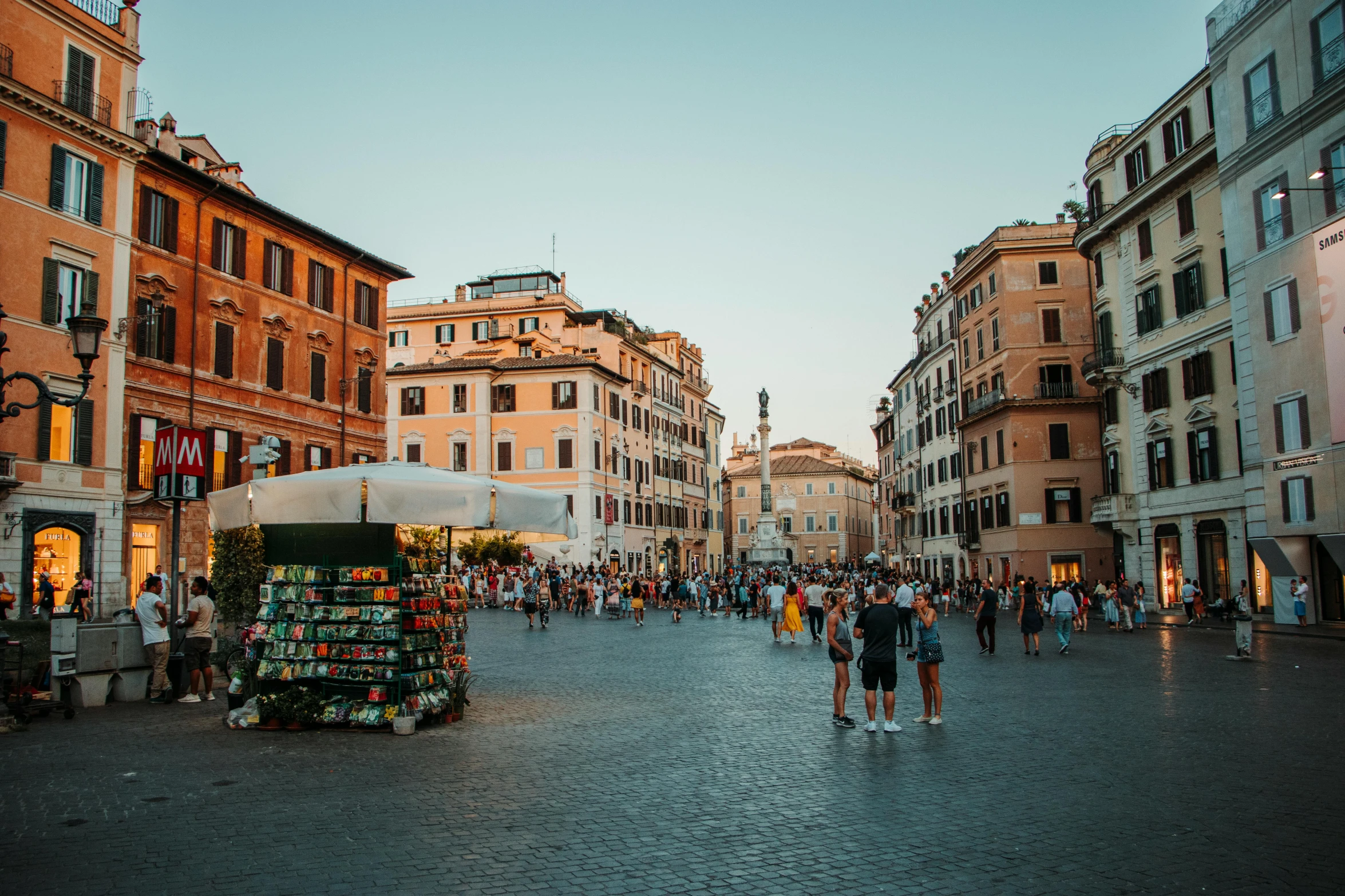 a group of people walking down a street next to tall buildings, by Julia Pishtar, pexels contest winner, neoclassicism, colosseo, town square, summer evening, sunken square