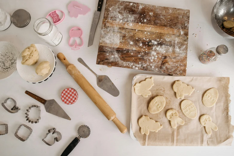 a table topped with a cutting board covered in cookies, by Sylvia Wishart, unsplash, folk art, metal kitchen utensils, detailed product image, various posed, dough sculpture