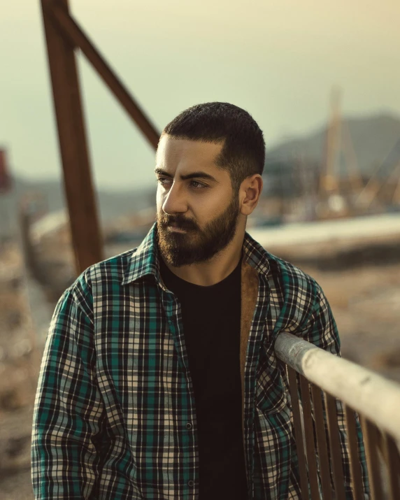 a man with a beard standing on a bridge, a colorized photo, by Ismail Acar, trending on pexels, non binary model, middle eastern skin, wearing a plaid shirt, focused stare