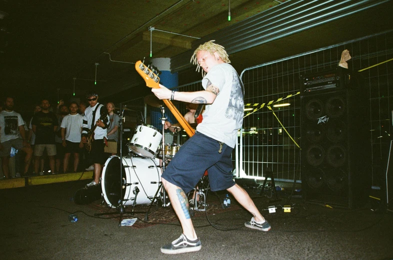 a man standing on a stage with a guitar, unsplash, lowbrow, at a skate park, extremely pale blond hair, wielding a crowbar, kombi