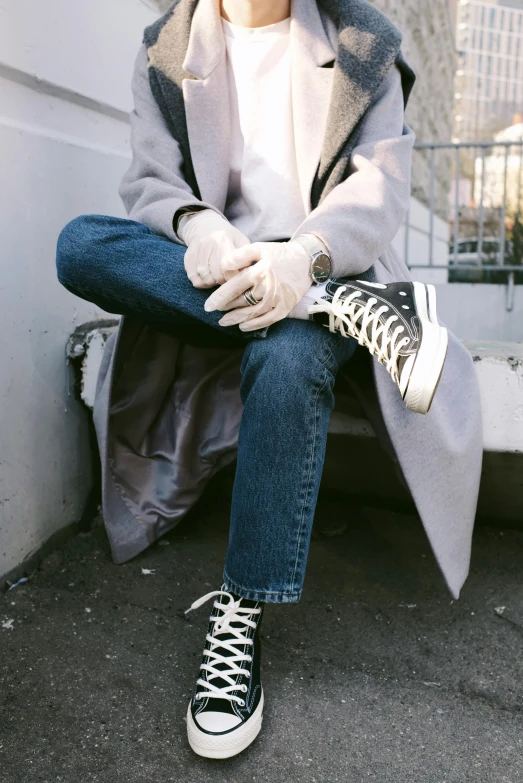 a woman sitting on a ledge with her legs crossed, inspired by jeonseok lee, trending on pexels, blue jeans and grey sneakers, wearing a long coat, pastel goth aesthetic, androgynous male