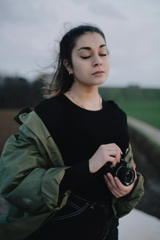 a woman standing on the side of a road holding a camera, wearing dark green bomber jacket, portrait featured on unsplash, in a field, portrait sophie mudd