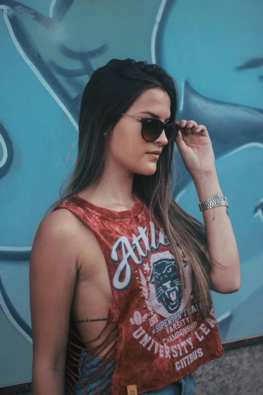 a woman standing in front of a graffiti wall, an airbrush painting, inspired by Elsa Bleda, trending on pexels, wearing red tank top, aviators, long ashy hair, tanned skintone