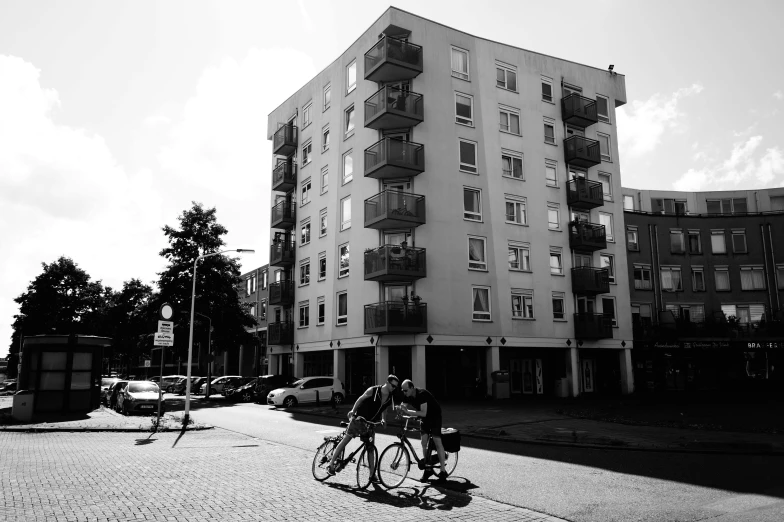 a couple of people riding bikes down a street, a black and white photo, by Jacob Toorenvliet, concrete housing, full building, uploaded, high quality upload