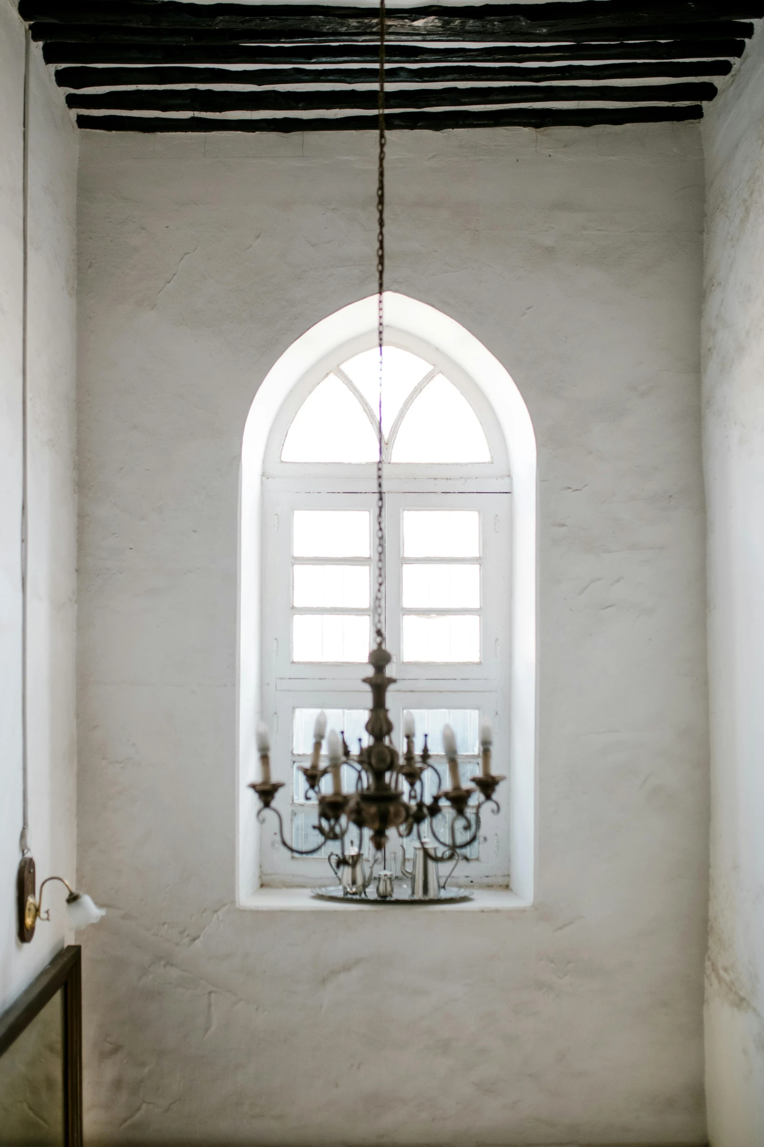 a room with a chandelier and a window, inspired by Riad Beyrouti, trending on unsplash, baroque, white wall, gothic arch frame, rustic and weathered, gallery lighting