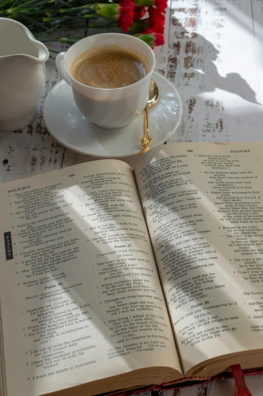 an open book sitting on top of a table next to a cup of coffee, sun dappled, with full descriptions, holy