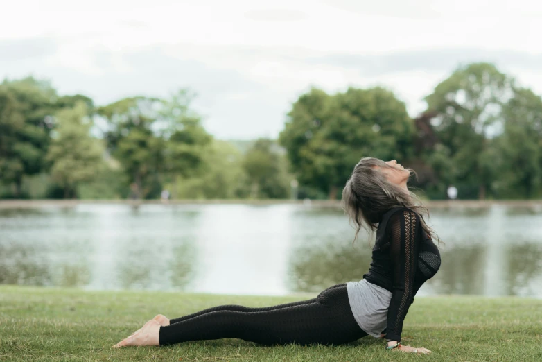 a woman doing yoga in front of a lake, by Rachel Reckitt, arabesque, in the park, profile image, looking towards camera, grey