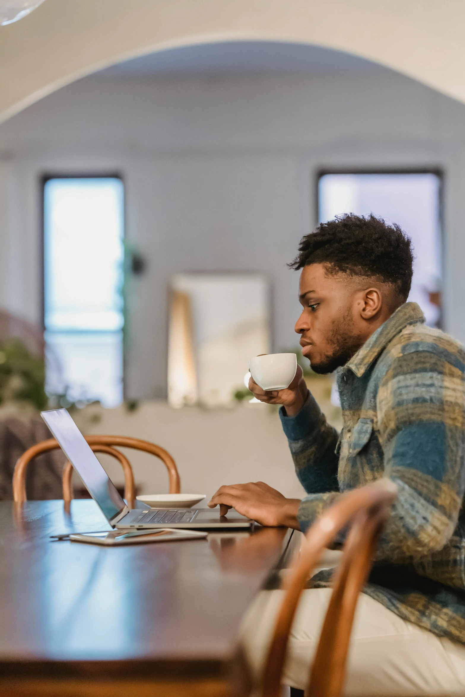 a man sitting at a table using a laptop, trending on pexels, next to a cup, man is with black skin, looking left, open plan