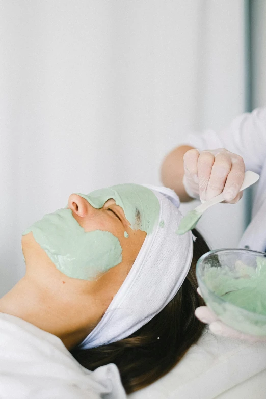 a woman getting a facial mask at a spa, by Nicolette Macnamara, pastel green, seafoam green, coerent face and body, manuka