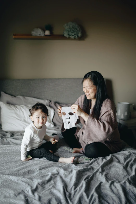 a woman sitting on top of a bed next to a child, inspired by Li Di, pexels contest winner, wearing giant paper masks, manuka, 2 years old, grey