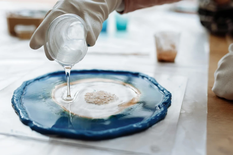 a person in white gloves pouring something into a blue plate, a hyperrealistic painting, inspired by Vija Celmins, trending on unsplash, chemical substances, geode, silicone skin, cast glass