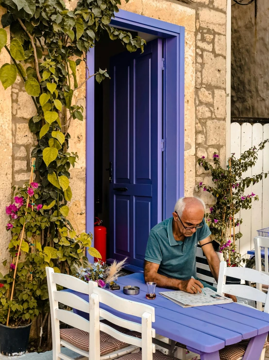 a man sitting at a table in front of a blue door, delightful surroundings, greek setting, old man doing hard work, 2019 trending photo