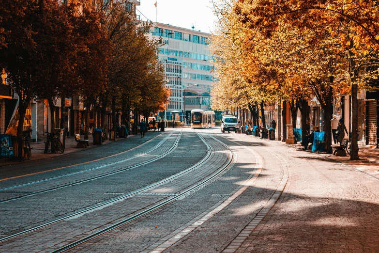 a train traveling down a city street next to tall buildings, pexels contest winner, autumn leaves on the ground, vallejo, thumbnail, street tram