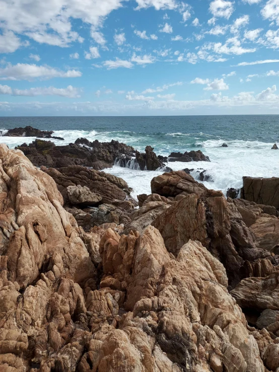 a man standing on top of a rock next to the ocean, by Hubert van Ravesteyn, panoramic widescreen view, boulders, layers of strata, brown