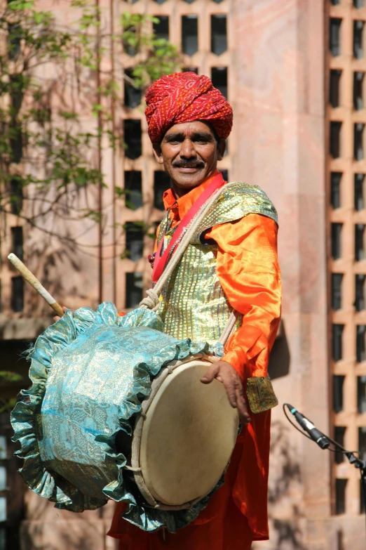 a man in a turban holding a drum, dressed in a jodhpuri suit, on a bright day, wearing a fancy dress, surrounding the city