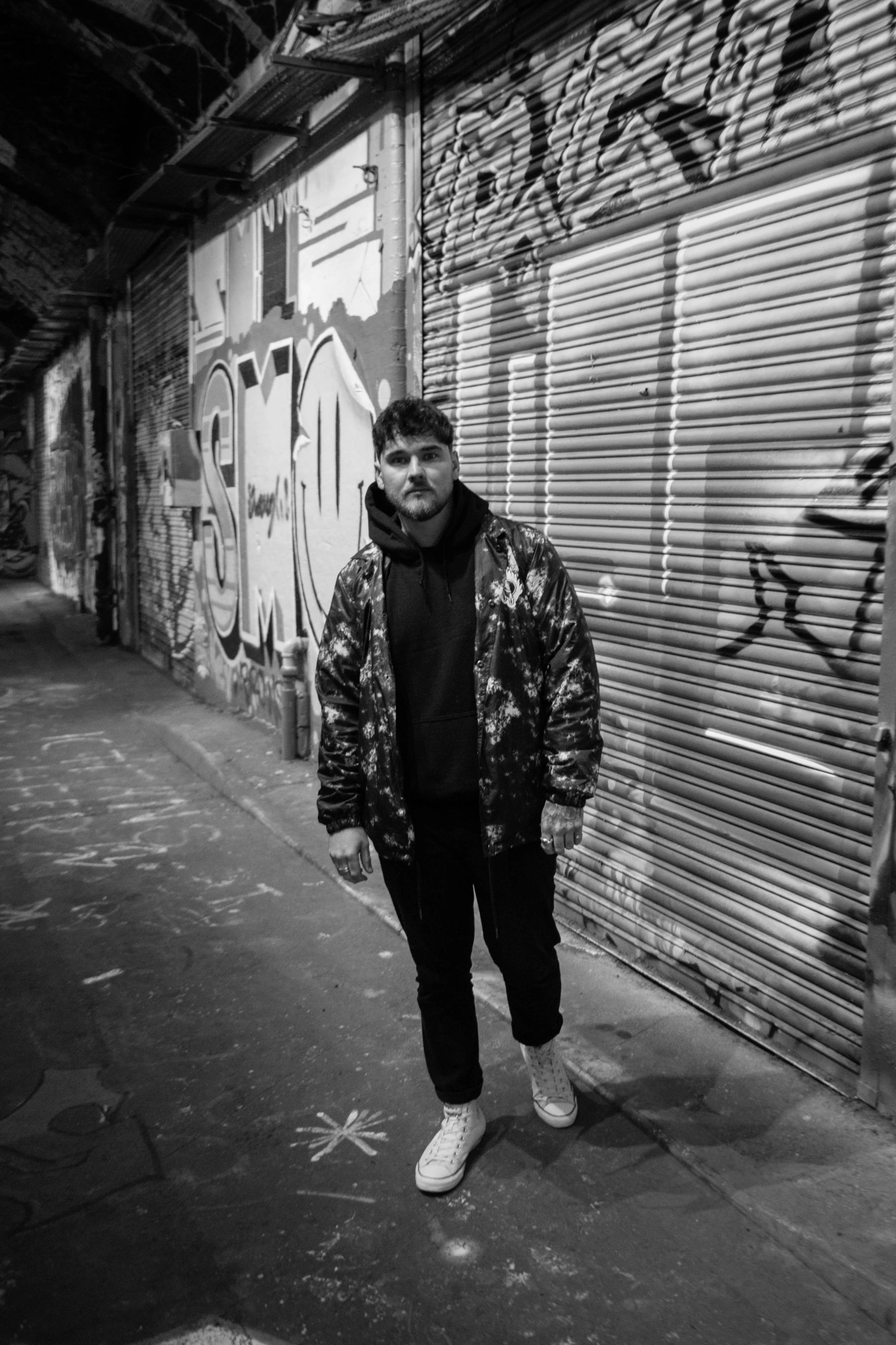 a man standing in front of a graffiti covered wall, a black and white photo, inspired by Ion Andreescu, charli bowater and artgeem, evening time, wearing camo, wearing a baggy