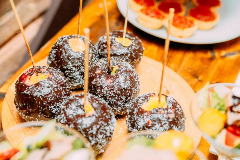 a wooden table topped with donuts covered in powdered sugar, by Julia Pishtar, unsplash, candy apple, skewer, ?black apples, square