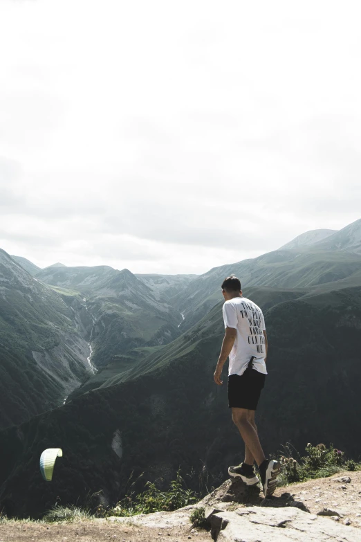 a man standing on top of a mountain holding a tennis racquet, by Jacob Toorenvliet, happening, looking down at the valley, wearing shorts and t shirt, trending photo, facing away