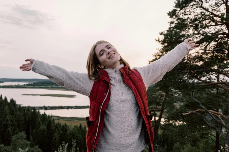 a woman standing on top of a lush green hillside, pexels contest winner, happening, greta thunberg smiling, wearing a red gilet, pose(arms up + happy), evening time