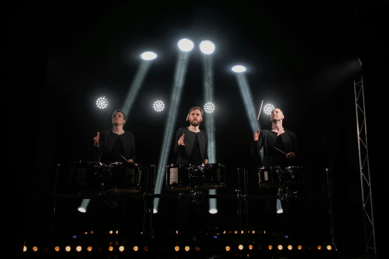 a group of people standing on top of a stage, an album cover, pexels, bauhaus, drum kit, glowing with silver light, trio, sleek hands