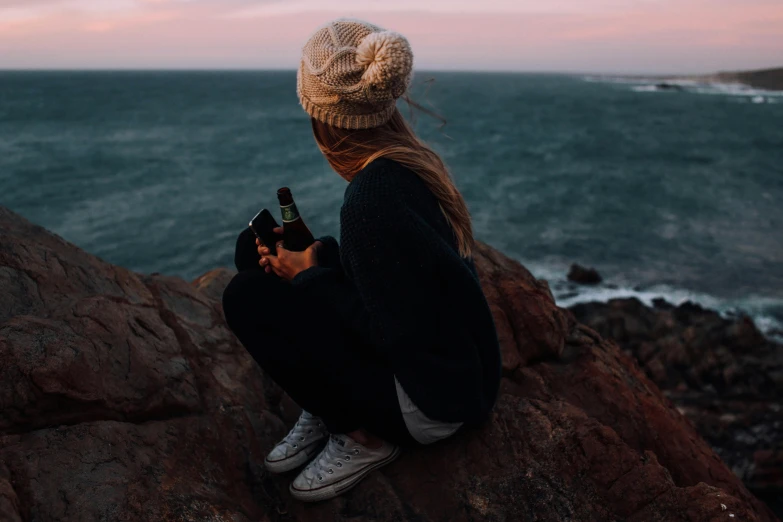 a woman sitting on top of a rock next to the ocean, trending on pexels, realism, holding a bottle of beer, moody aesthetic, profile image, vapourwave