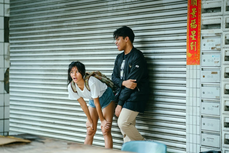 a man and a woman standing next to each other, by Tan Ting-pho, trending on pexels, graffiti, teenage boy, embarrassing, asian female, sitting on the ground