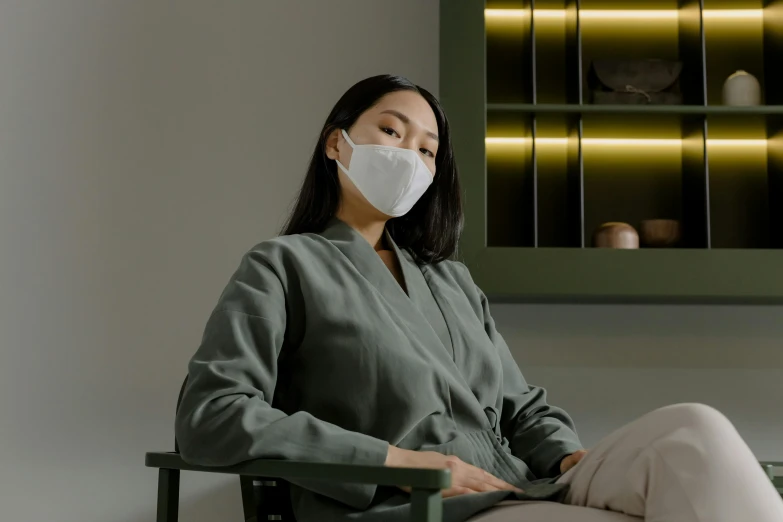 a woman sitting in a chair wearing a face mask, inspired by Fei Danxu, trending on pexels, sōsaku hanga, wearing nanotech honeycomb robe, (mist), green facemask, ignant