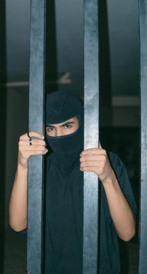 a man with a black face covering his face behind bars, an album cover, inspired by Bernardino Mei, trending on pexels, hurufiyya, ninja outfit, malaysian, lgbtq, 2 0 0 0's photo