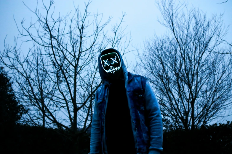 a person wearing a mask with trees in the background, an album cover, pexels contest winner, graffiti, moody blue lighting, black hood, backlit glow, bleak lighting