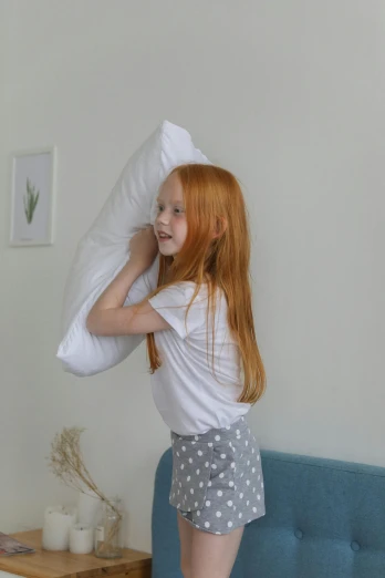 a little girl standing on top of a bed holding a pillow, flowing ginger hair, full product shot, dressed in a white t shirt, right hand side profile