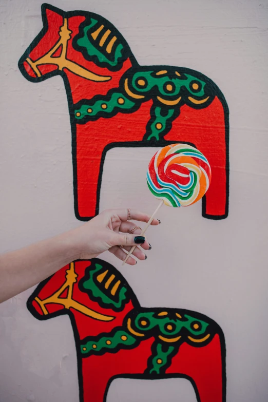 a person holding a lollipop in front of a painted horse, by Julia Pishtar, trending on pexels, toyism, on a wall, sweets, 15081959 21121991 01012000 4k