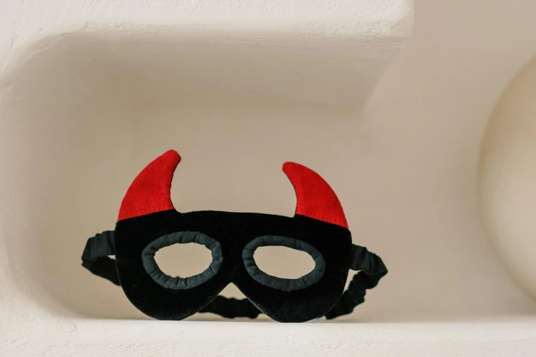 a black mask with red horns on a shelf, by Emma Andijewska, pexels, children's toy, velvet, harness, high angle close up shot