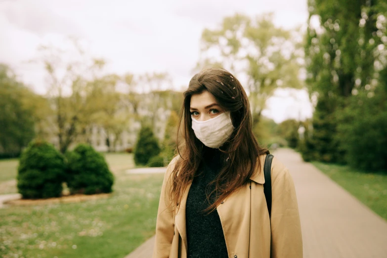 a woman wearing a face mask in a park, by Emma Andijewska, trending on pexels, renaissance, light brown coat, polluted, 256435456k film, people outside walk