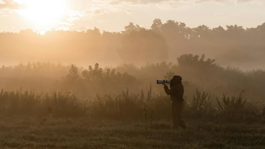 a person standing in a field with a camera, a picture, by Adam Marczyński, tonalism, 8k hdr morning light, gunfire, wildlife photograph, summer morning