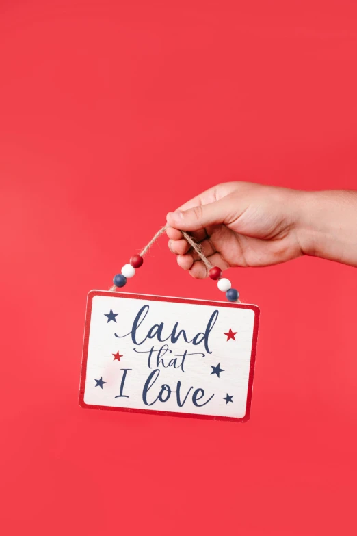 a hand holding a sign that says land with love, by Julia Pishtar, shutterstock, folk art, red white background, full product shot, ornament, 4th of july